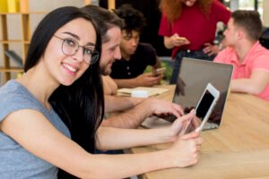 portrait-smiling-woman-holding-digital-tablet-looking-camera-while-sitting-beside-her-friends-using-electronic-gadgets-book-wooden-desk (2)